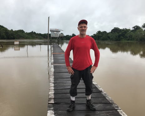 Dom Phillips stands on a wooden jetty over a wide river, rainforest and a stilted hut behind him