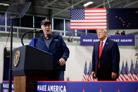a man speaks into a microphone as Donald Trump stands behind him